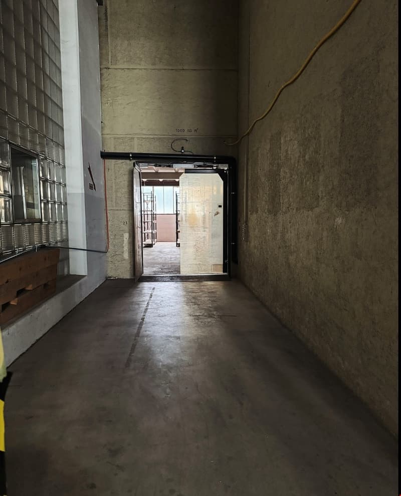 Empty corridor with gray concrete floor, walls, visible metal piping, single open door.