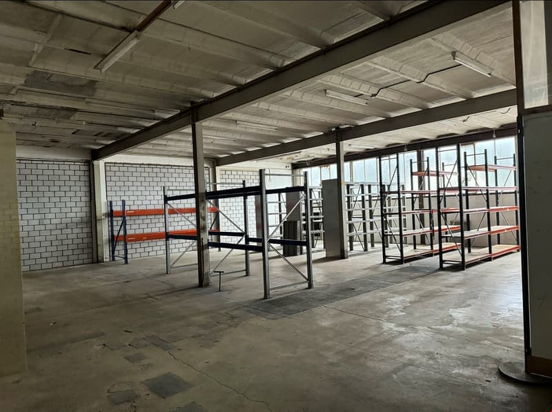 Empty warehouse with metal shelves, metal columns, white brick walls, concrete floor, exposed ceiling beams, large windows, metal stairs.