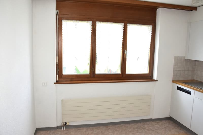 Empty room with white walls, wooden window frame, white radiator, tiled backsplash, white cabinets, and a sink.