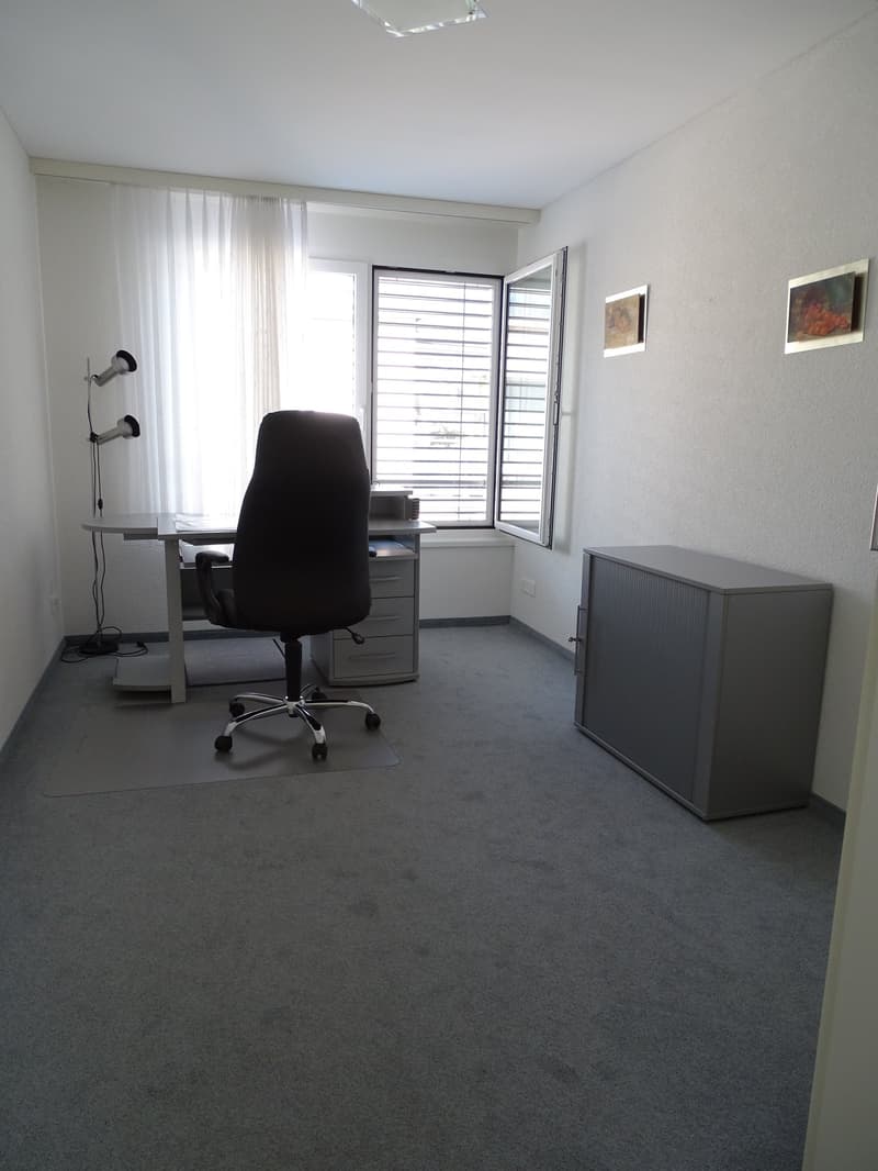 The office room is equipped with a desk, a chair, and a lamp. There is also a shelf with books and a power outlet. There are two windows with blinds and two picture frames on the wall. The room has a tiled floor and a ceiling with a light fixture.