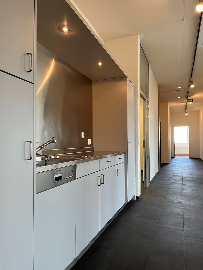 Modern kitchen with white cabinets, stainless steel appliances including a dishwasher, and dark tile flooring. The kitchen leads into a hallway with additional rooms.