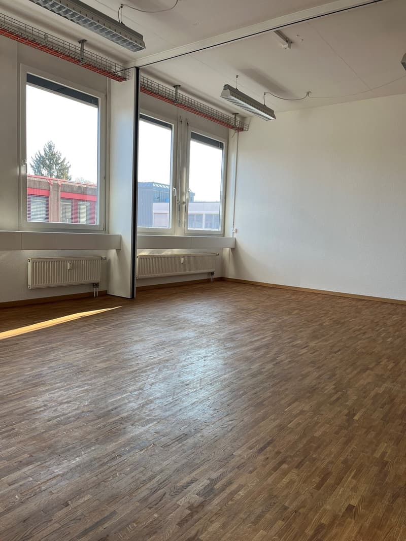 Large open room with hardwood flooring, white walls, and large windows providing natural light. The room has ceiling-mounted heating and cooling units, and shelving units along the walls.