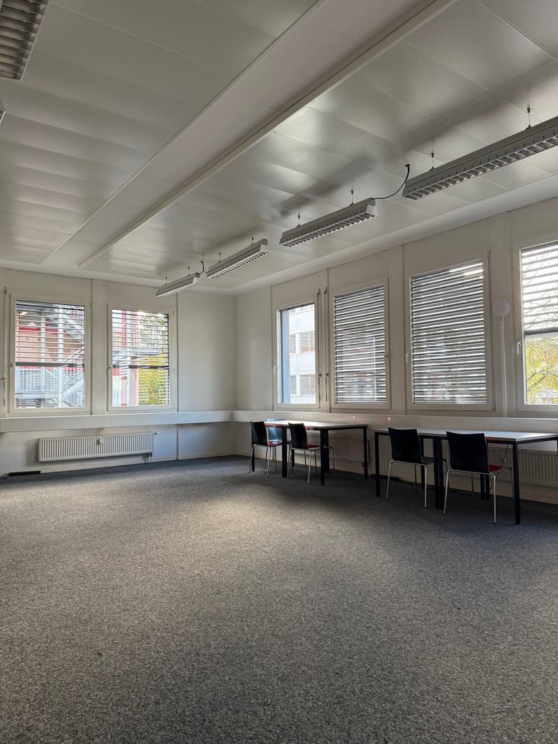 Empty meeting room with grey carpet, tables, chairs, windows, radiators, and ceiling lamps.
