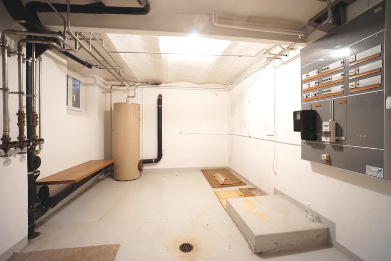 A utility room with wooden bench, large water heater, power boxes, and pipes on the walls.