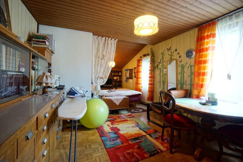 Room with bed, wooden cabinet, carpet, table with chairs, lamp, decorative ball, ironing board, mirror, clock, shelves, window with curtains