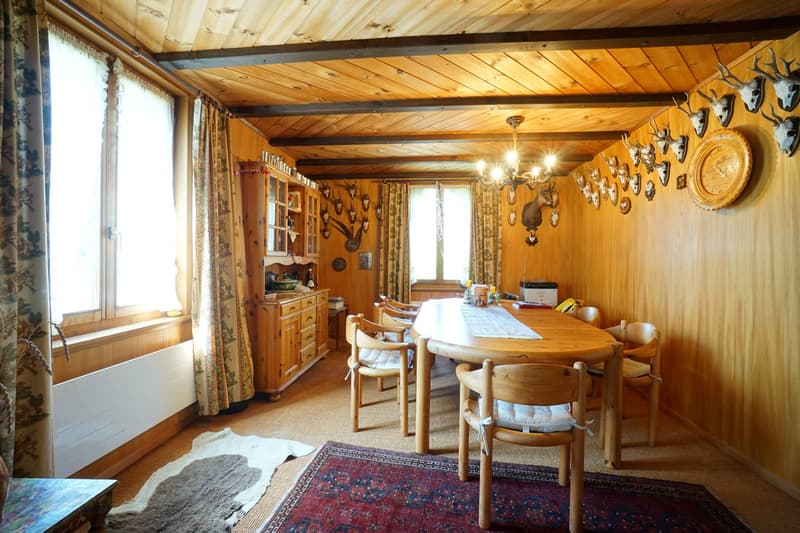 Dining room with wooden dining table, wooden chairs, wooden cabinet, wall with deer trophies and mounted clock, wooden flooring, radiator, wooden ceiling, and two windows with curtains