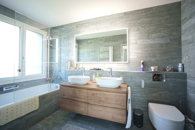 modern bathroom with gray tiles, wooden vanity, two sinks, large mirror, bathtub, toilet, and shower area