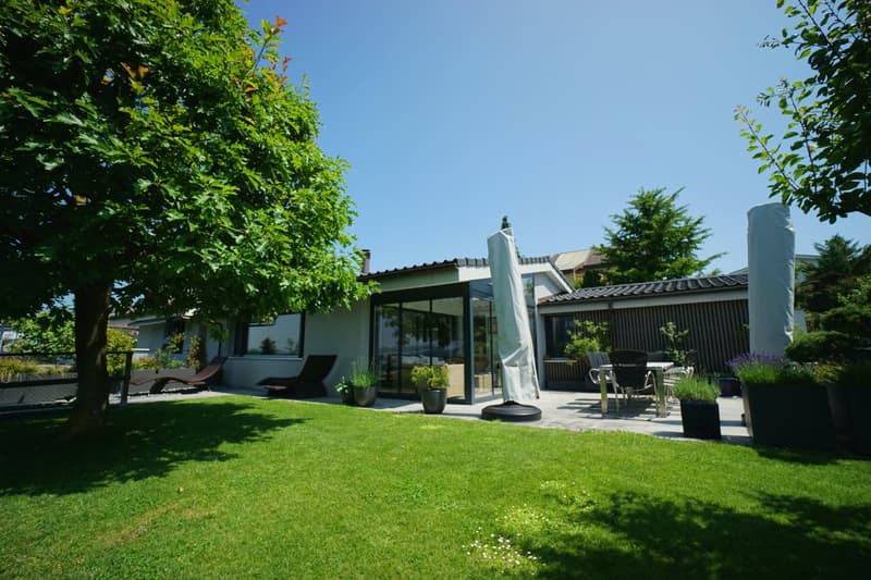 well-maintained garden with green lawn, patio with table and chairs, lounge chairs, potted plants, and trees