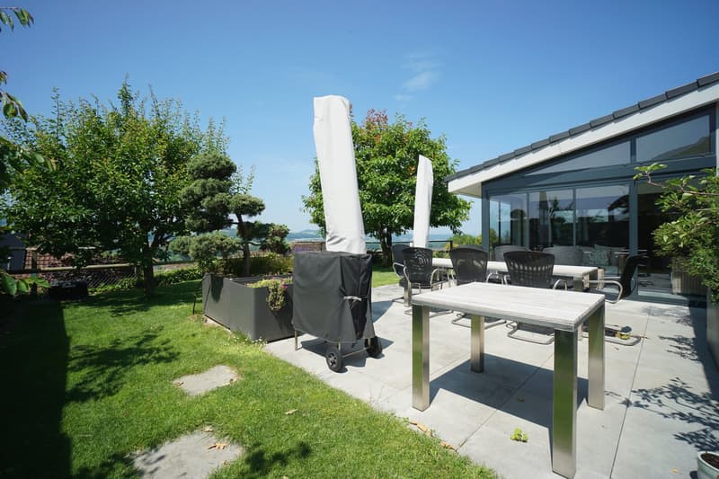 Outdoor dining area with table and chairs, covered by umbrellas, covered patio, garden with trees and plants, view of the mountains