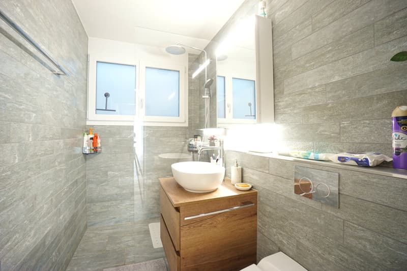 Bathroom with a white sink, wooden cabinet, shower, and tiled walls.