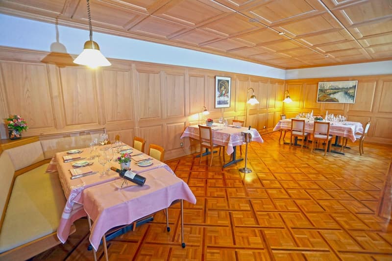 Spacious dining room with wooden paneled walls and ceiling, parquet flooring, multiple dining tables set with tablecloths and place settings, hanging light fixtures, and a framed artwork on the wall.