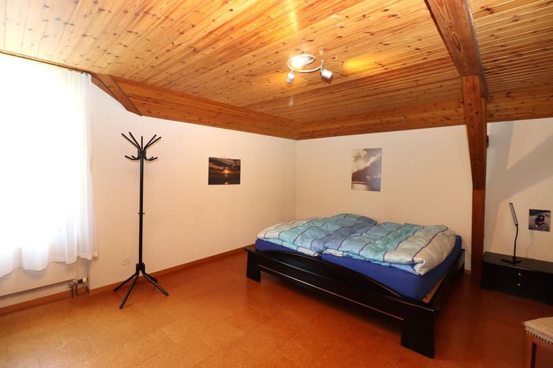 Spacious bedroom with wooden ceiling, white walls, a bed with a blue and green comforter, a coat rack, and framed artwork on the walls.