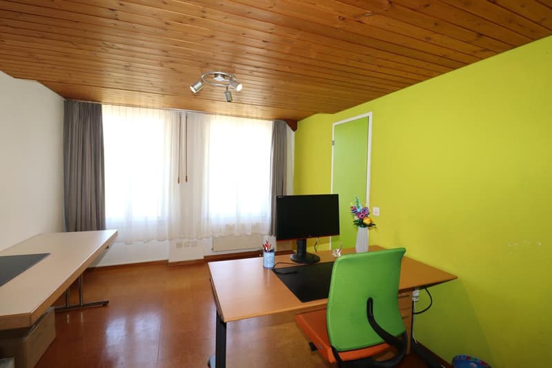Spacious home office with wooden ceiling, large windows, desk, green office chair, and a TV mounted on the wall