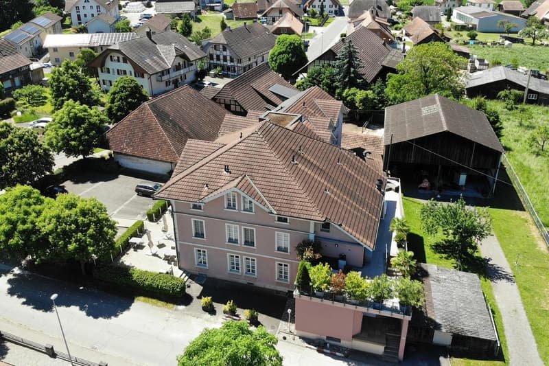Aerial view of a picturesque village with traditional Swiss-style buildings, red tile roofs, and lush greenery surrounding the structures. There appears to be a parking area visible in the image.