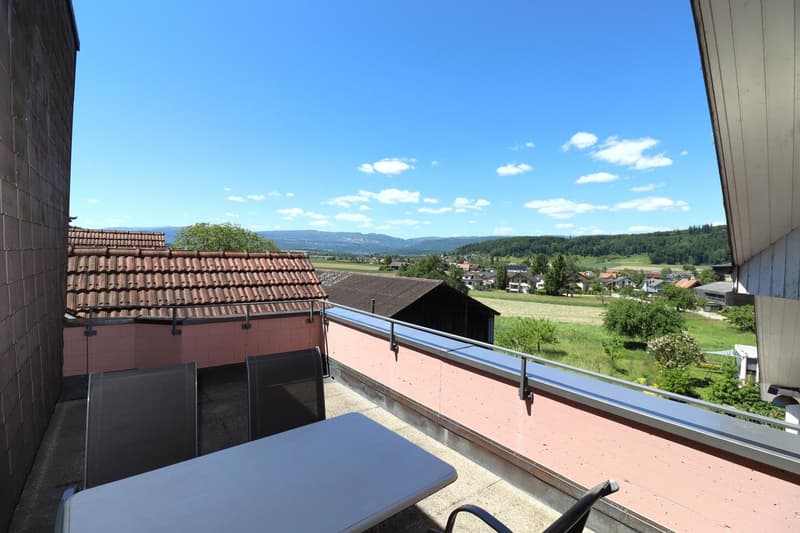 Balcony with a view of the countryside, including rooftops, trees, and distant hills. The balcony has a railing and appears to be part of a larger building.