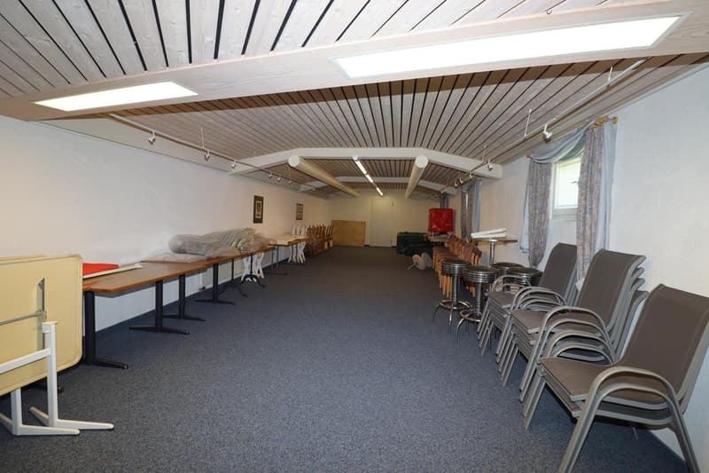 The image shows a large open space with a high ceiling and wooden paneled walls. The room is equipped with several long tables and chairs, suggesting it is used for meetings, events, or gatherings. The space appears to be well-lit with track lighting and