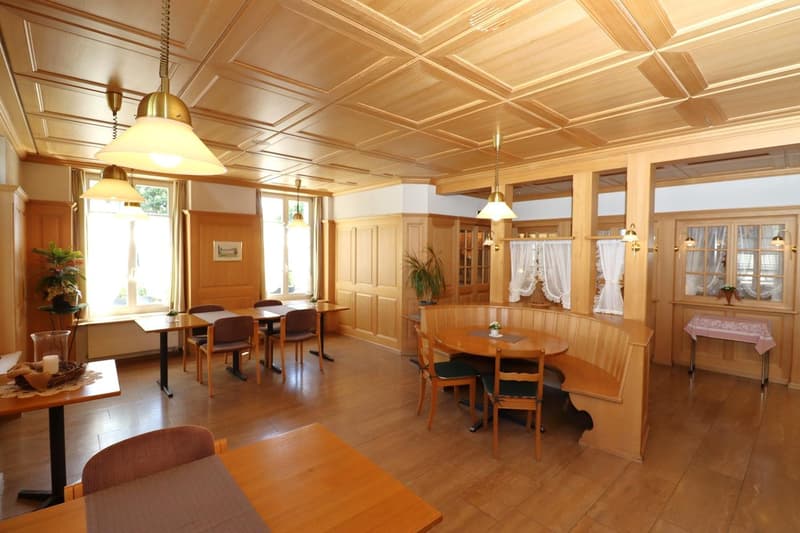 Spacious dining room with wooden paneled walls and ceiling, large windows, wooden tables and chairs, fireplace, and decorative elements like plants and curtains.