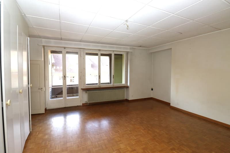 Spacious living room with large windows, hardwood flooring, and a radiator. The room appears to be in need of some renovation or updating.