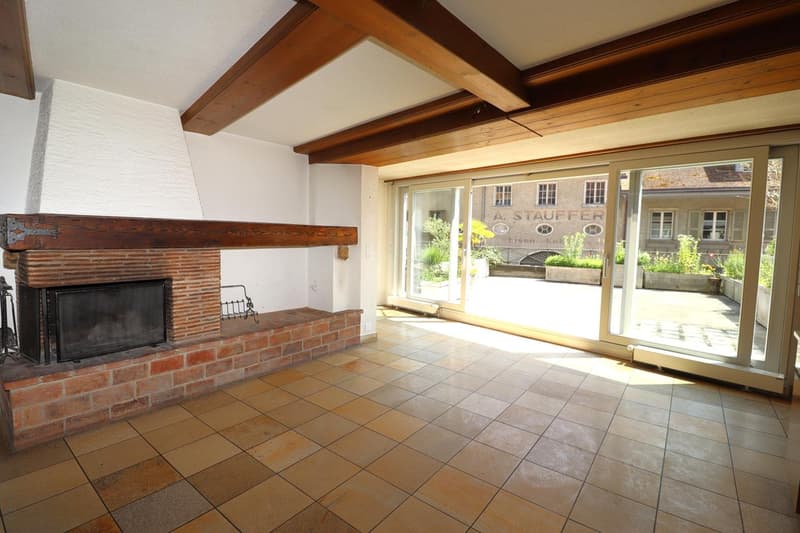 Spacious living room with exposed wooden beams, a brick fireplace, and large windows overlooking an outdoor area. The room has tiled flooring and a cozy, rustic atmosphere.