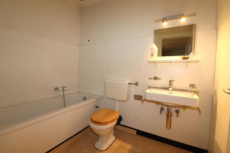 The image shows a bathroom with a bathtub, toilet, and a vanity with a sink. The walls are white, and the floor appears to be hardwood. There is a shelf above the sink, and the lighting fixtures are visible on the wall. The bathroom also has a private was