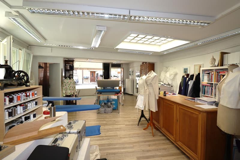 This image shows a sewing/tailoring workshop with various sewing machines, fabrics, and other equipment. The room has a large open layout with wooden floors and shelves along the walls. There is a private washing machine visible, indicating that this spac