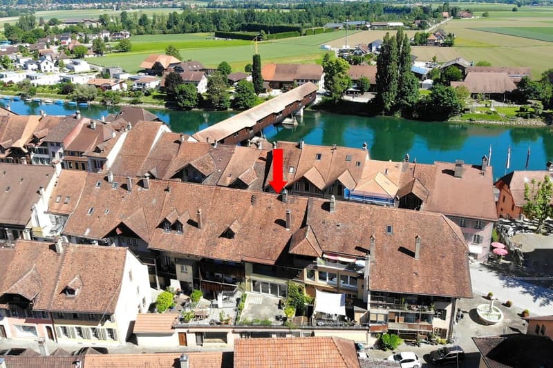 Aerial view of a historic town with traditional Swiss-style buildings, a covered bridge over a river, and surrounded by lush green fields and forests.