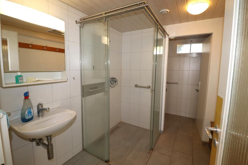 Bathroom with a white sink, glass shower enclosure, white tiles, mirror, and bottle of cleaning product.