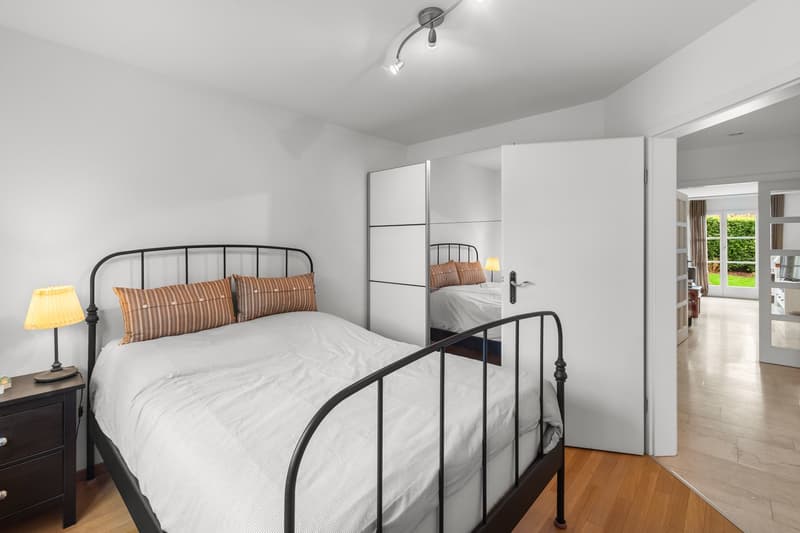white bed, white sheets, dark brown nightstand with lamp, white walls, glass door to hallway, bedroom with a bed reflected in a mirrored wall