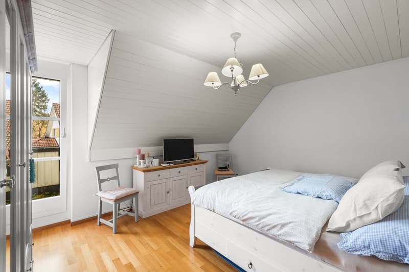 Bedroom with wooden floor, white walls, large bed with white and blue pillows, white nightstand, chair, desk, monitor, lamp, glass door with view of exterior
