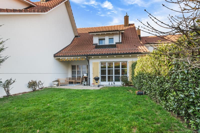 Two-story house with a tiled roof, white walls, wooden porch, large windows, well-maintained lawn, and a garden with shrubs and a barbecue.