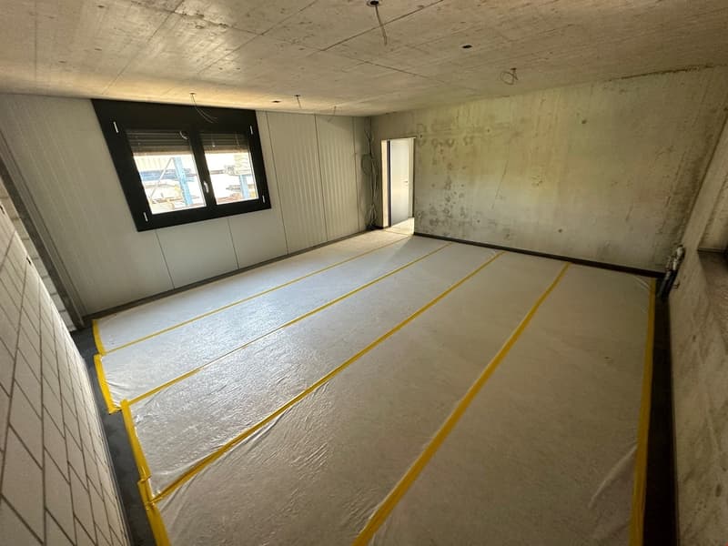 Unfinished room with concrete walls and floor, black window frame, and yellow and gray flooring material covering the floor.