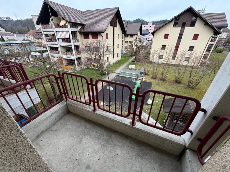 Red railings on balcony, view of the green grass, small white playhouse, a slide, other buildings in the background