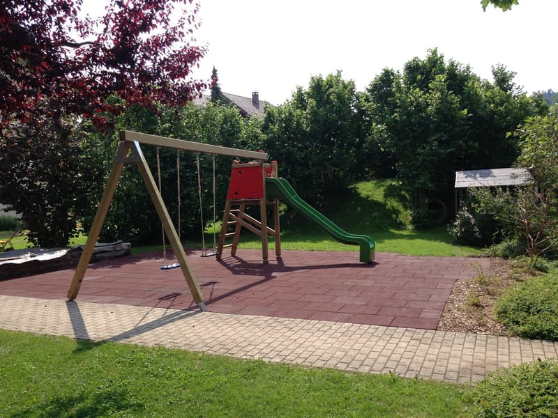 outdoor playground with swing, slide, brick ground, surrounded by grass and trees