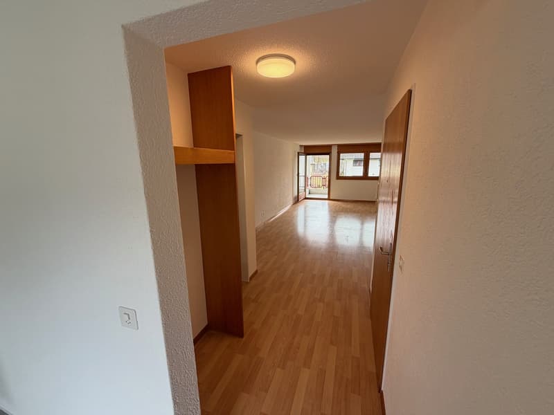 A hallway with wooden floors and white walls. A light is on the ceiling. There is a wooden wardrobe on the left and a door at the end of the hallway.