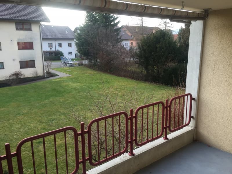Balcony with red railings, overlooking a grassy field and trees, with a building in the background