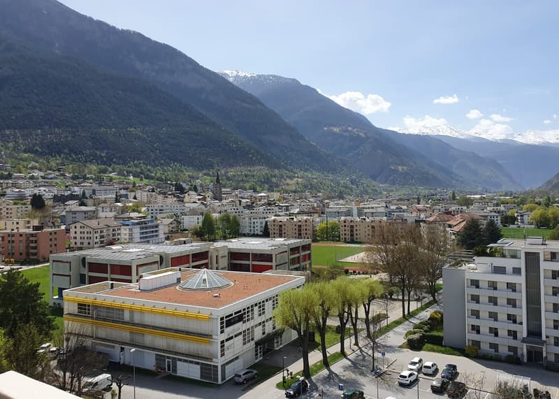 Aerial view of a building with a large roof, surrounded by trees and parked cars, with a mountain range in the background.