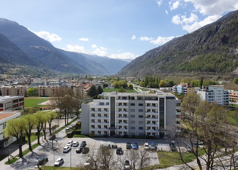 Aerial view of a multi-story building with a parking lot in front, surrounded by trees and mountains in the background.