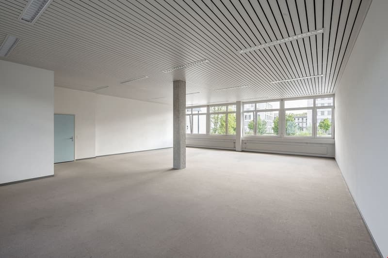 Large open office space with high ceilings, white walls, and large windows providing a view of the surrounding area. The space has a clean, modern design with a suspended ceiling and exposed beams.