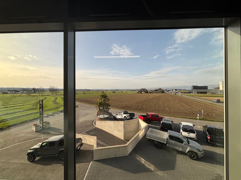 View of a parking lot with several pickup trucks and SUVs parked, a concrete structure, and a field in the background.