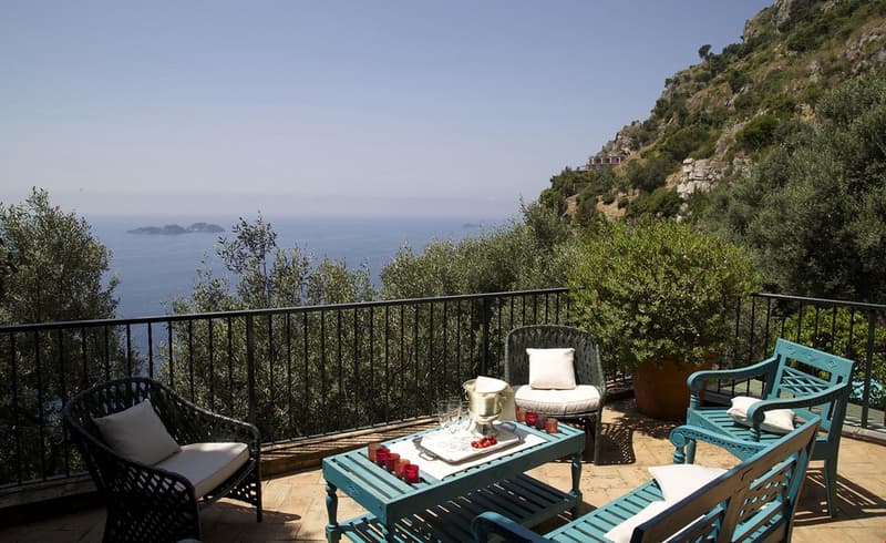 The image shows an outdoor terrace or balcony with a stunning view of the ocean and surrounding cliffs. The terrace is furnished with comfortable patio furniture, including chairs, a table, and decorative elements like pillows and a tray with glasses. The
