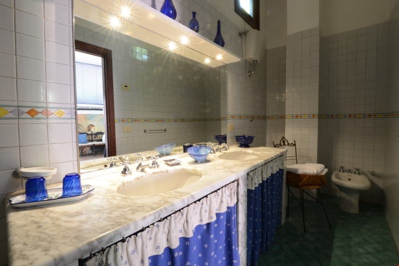 Tiled bathroom with marble countertop, sinks, and decorative blue and white accents. Shelves with blue vases and dishwasher visible.