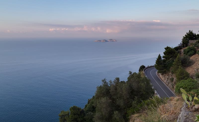 The image shows a scenic coastal landscape with a winding road along the cliffside overlooking the ocean. In the distance, a small island can be seen on the horizon. The sky is filled with soft, pastel-colored clouds at sunset. The surrounding vegetation