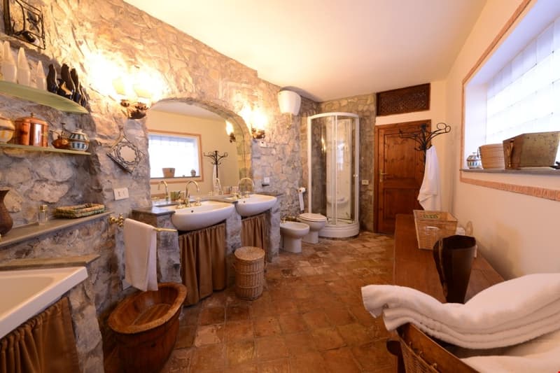 Large bathroom with stone walls, double vanity sinks, shower enclosure, wooden shelves, and rustic decor elements like baskets and towels