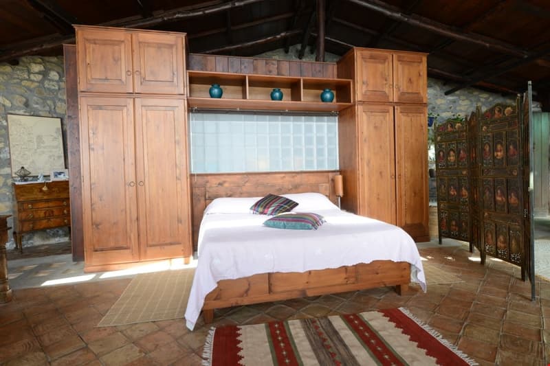 Rustic bedroom with wooden furniture, including a bed, armoire, and shelving units. The room has a stone wall and exposed wooden beams in the ceiling. There is also a decorative screen or room divider visible in the background.