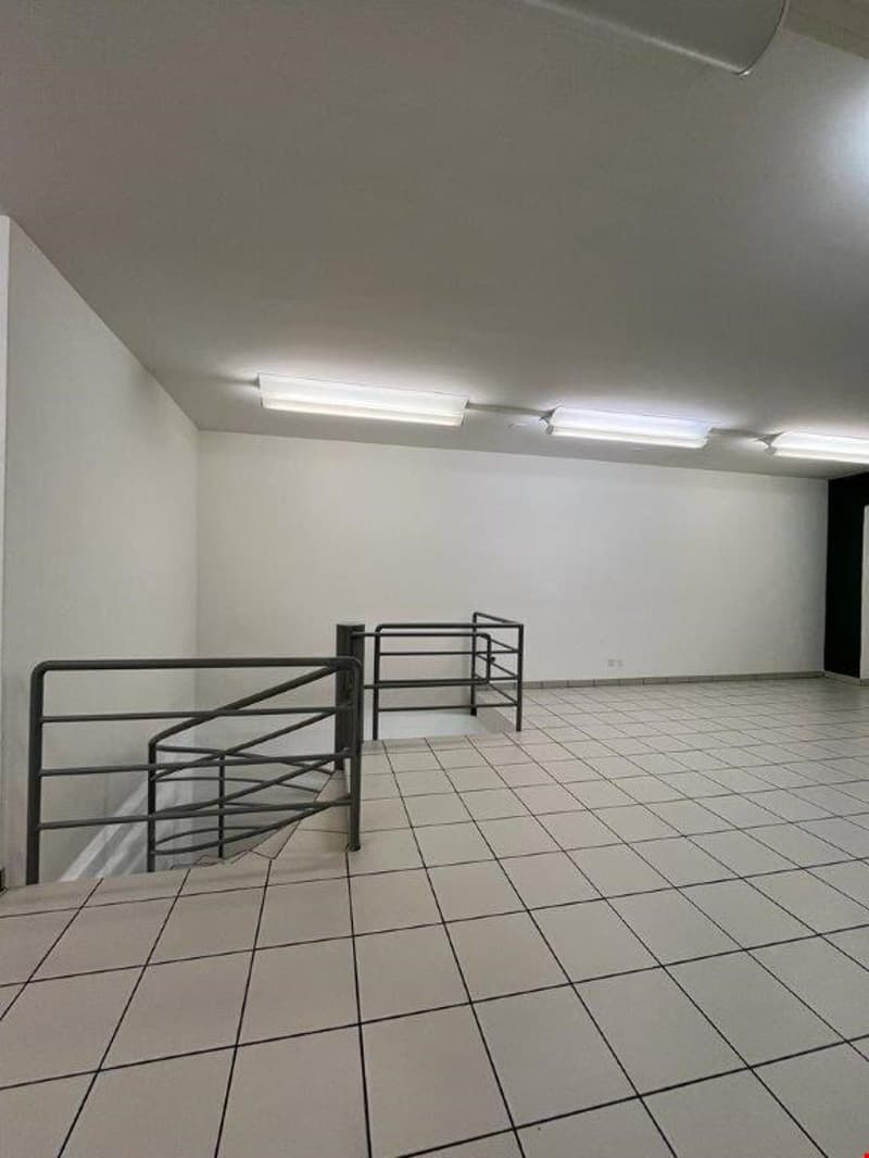 Empty room with white walls, tiled floor, and overhead lighting. The room appears to be a storage or utility space, with metal shelving units along the walls.