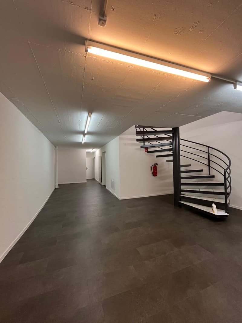 The image shows a long hallway with a spiral staircase in the center. The walls are painted white and the floor is a dark concrete material. The hallway is illuminated by long fluorescent light fixtures on the ceiling.