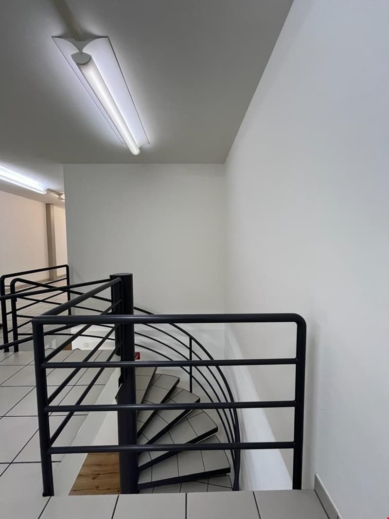 The image shows a spiral staircase with a black metal frame. The stairs appear to be made of wood or a similar material. The staircase is located in a room with white walls and a ceiling light fixture.
