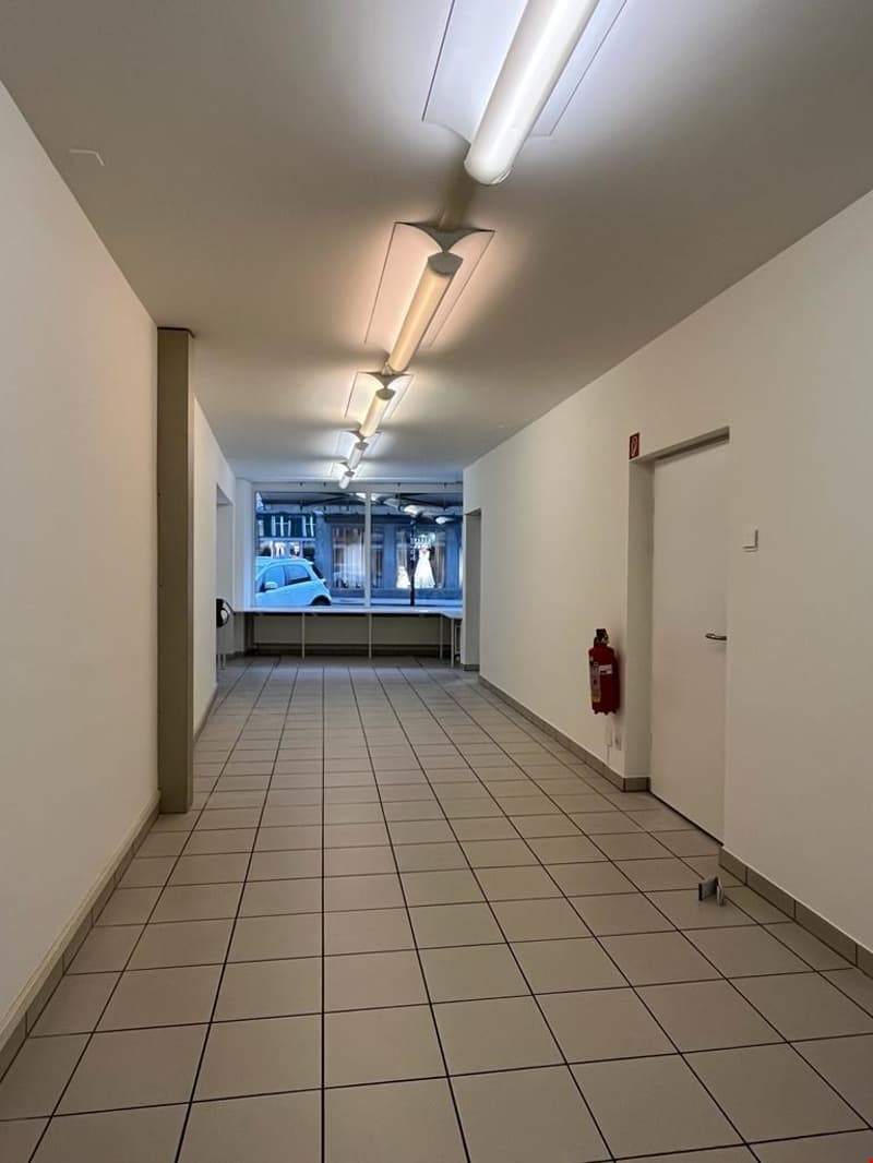 Long, tiled hallway with fluorescent lighting fixtures overhead. The hallway leads to a window at the end, providing a view of the exterior. A fire extinguisher is mounted on the wall.