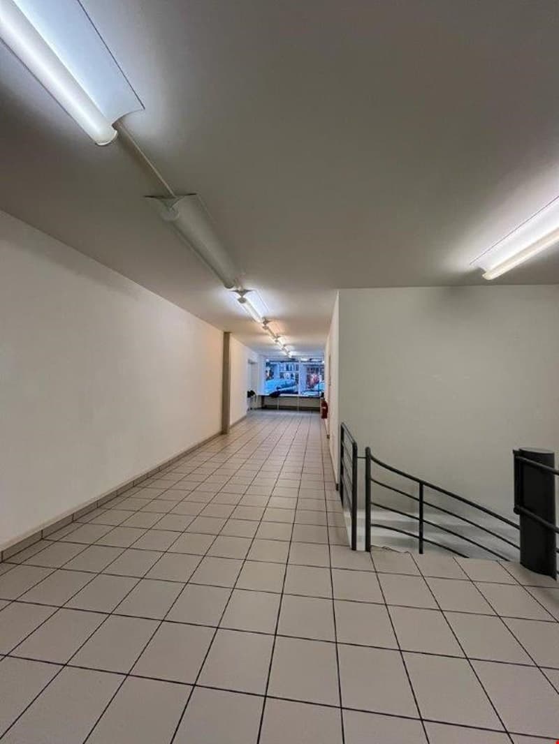 Long hallway with tile flooring, white walls, and overhead lighting fixtures. The hallway appears to lead to another room or area at the end.