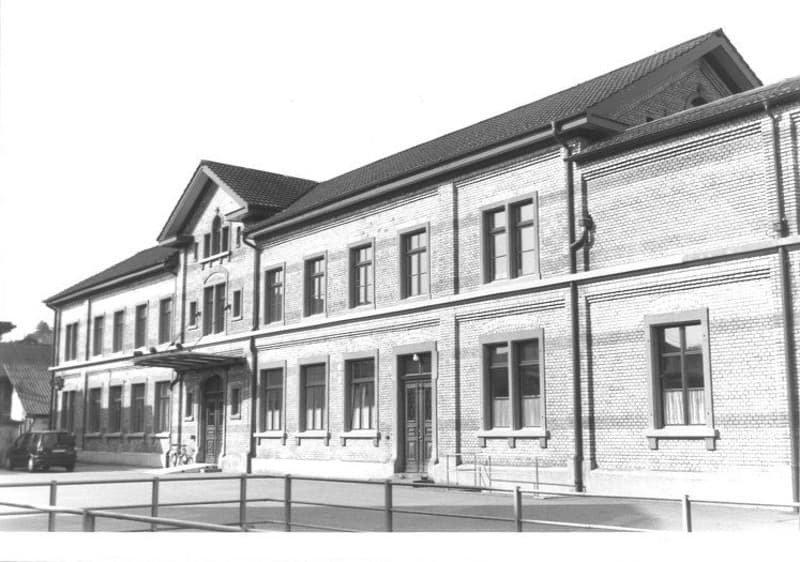 two story brick building, many windows, roof has a steeple, fence in front, bicycles leaning against building, car parked to the left